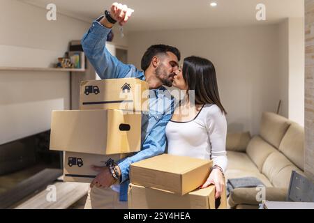 Ein junges Paar küsst sich, während es Pappkartons und Schlüssel für ihre neue Wohnung hält, und feiert den Zusammenzug Stockfoto