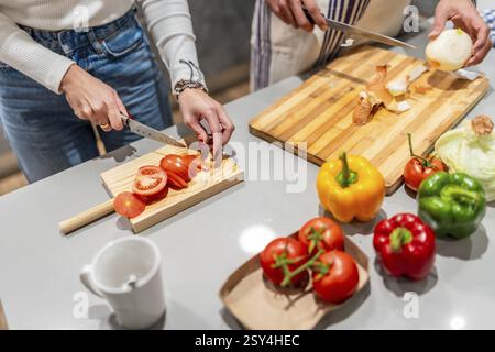 Zwei Köche schneiden in einer modernen Küche frische Tomaten und Zwiebeln auf hölzernen Schneidebrettern, umgeben von farbenfrohen Paprika Stockfoto