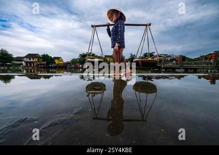 Ein Obstverkäufer mit konischem Hut spaziert entlang des Flusses Thu Bon, Sông Thu Bồn, der sich im Wasser spiegelt und Obstkörbe trägt. Stockfoto