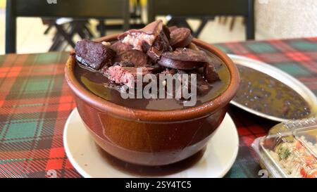 Dampfende brasilianische Feijoada, ein Eintopf aus schwarzen Bohnen mit verschiedenen Fleischgerichten, serviert in einem traditionellen Tontopf Stockfoto