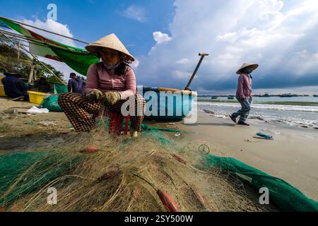 Eine Frau mit konischem Hut sitzt am Strand von Mui ne, Mũi Né und entwirrt Fischernetze, viele Fischerboote und Sturmwolken in der Ferne. Stockfoto