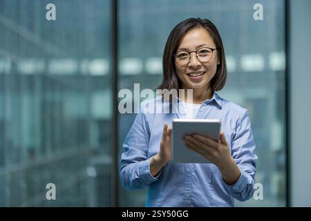 Lächelnde asiatische Geschäftsfrau, die ein Tablet vor einer Glasfassade hält und direkt in die Kamera blickt. Stockfoto