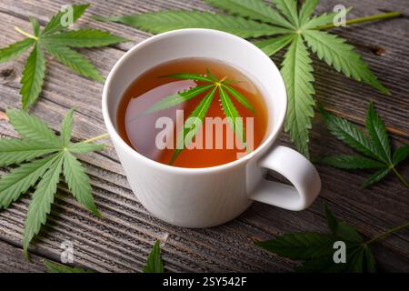 Weiße Tasse Cannabistee mit frischen grünen Marihuanablättern auf einem Holztisch. Nahaufnahme von medizinischem Kräutertee Stockfoto