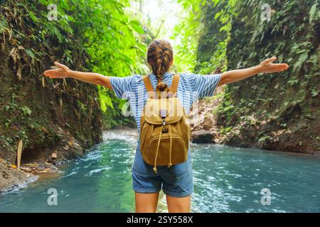 Weibliche Touristenin mit Rucksack und Blick auf eine fantastische Schlucht und einen Bergfluss. Frau mit erhobenen Armen im tropischen Wald. Märchenlandschaft. Stockfoto