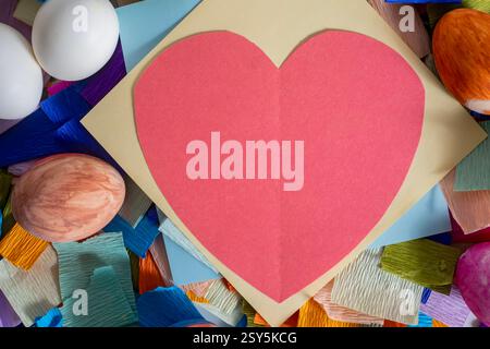 Ein Blick von oben auf ein rotes Herz, umgeben von bunten Bastelpapier und dekorierten Eiern, perfekt für Ostern oder saisonale Feierlichkeiten. Stockfoto