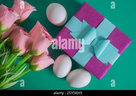 Blick von oben auf eine lila Geschenkbox mit blauem Band, rosa Rosen und weißen Eiern auf grünem Hintergrund. Stockfoto