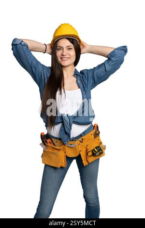 Eine lächelnde Bauarbeiterin mit gelbem Schutzhelm, Denim-Hemd und Werkzeuggürtel, die selbstbewusst mit den Händen hinter dem Kopf posiert. Th Stockfoto