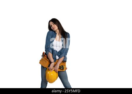 Eine junge Bauarbeiterin in einem Denim-Hemd, einem weißen Tanktop und einem Werkzeuggürtel, die einen gelben Schutzhelm mit besinnlichen Ausdrücken in der Hand hält Stockfoto