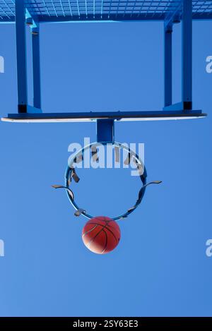 Ein Basketball läuft durch einen abgenutzten blauen Metallkorb vor einem klaren Himmel. Das Netz fehlt, zeigt eine städtische Outdoor-Sportanlage. Stockfoto