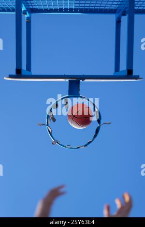Ein Basketball läuft durch einen abgenutzten blauen Metallkorb vor einem klaren Himmel. Das Netz fehlt, zeigt eine städtische Outdoor-Sportanlage. Stockfoto