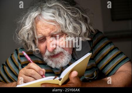 Ein älterer Mann mit lockigen grauen Haaren und Bart schreibt mit einem rosafarbenen Stift in ein Notizbuch, trägt ein gestreiftes Hemd und konzentriert sich zutiefst auf seine Gedanken. Stockfoto
