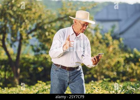 Mit Smartphone. Der Senior Mann steht tagsüber im Garten. Stockfoto