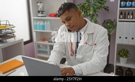 Junger hispanischer Mann, der in einem Krankenhauszimmer arbeitet, einen weißen Mantel und ein Stethoskop trägt, einen Laptop benutzt und am Telefon spricht Stockfoto