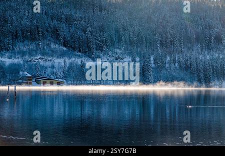 Mystischer Wintermorgen: Nebeliger See mit Waldspiegelung und schneebedeckten Bäumen Stockfoto