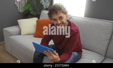 Ein junger Mann, der drinnen mit einem Tablet und einem Lächeln in einem modernen Wohnzimmer sitzt, bietet Komfort und Technologie in einer intimen Wohnumgebung. Stockfoto