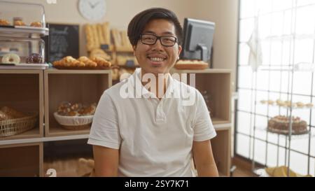Hübscher junger Mann, der drinnen in einer Bäckerei lächelt und im Hintergrund Gebäck und Brot serviert Stockfoto