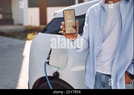 Der Ladevorgang wird auf dem Smartphone angezeigt. Nahaufnahme eines Mannes mit seinem Elektroauto. Stockfoto