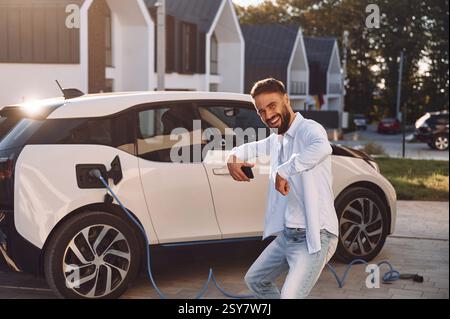 Lächeln und Spaß haben, während das Fahrzeug aufgeladen wird. Der junge, stilvolle Mann hat tagsüber ein Elektroauto. Stockfoto