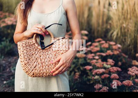Nahaufnahme einer stylischen Bio-Handtasche. Modische Frau, die im Sommer ein Kleid aus Leinen trägt und im Park eine Brieftasche aus Korb und eine Sonnenbrille hält. Stockfoto