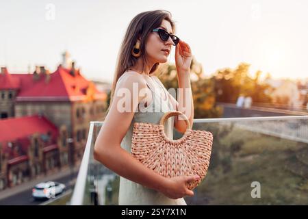 Porträt einer jungen modischen Frau, die abends Sommeroutfit auf der Stadtstraße trägt. Stilvolle Handtasche aus Strohrost. Weibliche Accessoires und Kleidung Stockfoto