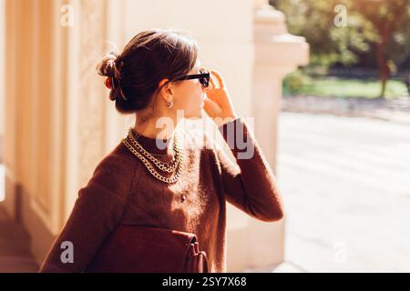 Modische Frau mit braunem Kaschmir-Pullover und gelbem Rock auf der Stadtstraße mit stilvoller Lederhandtasche kombiniert mit goldenem Schmuck und Stockfoto