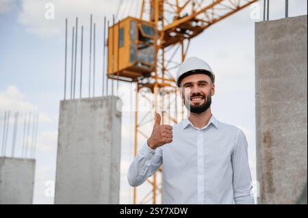 Lächelt und zeigt den Daumen hoch. Mann in Uniform arbeitet auf der Baustelle. Stockfoto