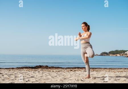 Ruhige Frau, die auf einem Bein steht und tief atmet und Stratching-Übungen macht und in den frühen Morgenstunden am Sandstrand mit ruhigen Meereswellen meditiert. Stockfoto
