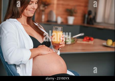Den Bauch berühren und das Glas mit Orangensaft halten. Schöne schwangere Frau ist zu Hause drinnen. Stockfoto