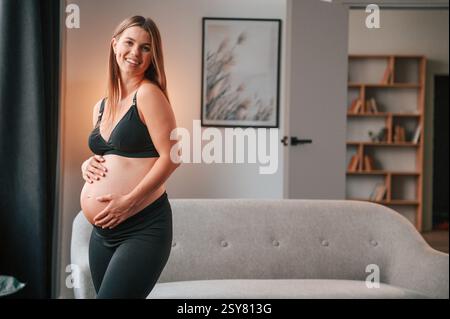Neben dem Sofa stehen und lächeln. Schöne schwangere Frau ist zu Hause drinnen. Stockfoto
