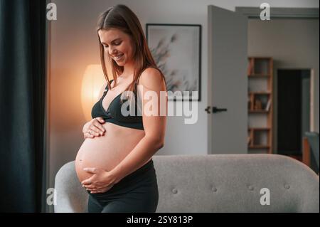 Neben dem Sofa stehen und lächeln. Schöne schwangere Frau ist zu Hause drinnen. Stockfoto