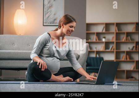 Yoga-Unterricht mit Laptop ansehen. Schöne schwangere Frau ist zu Hause drinnen. Stockfoto