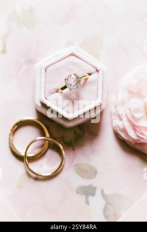 Ein atemberaubender Diamant-Verlobungsring befindet sich auf einer zarten Box, umgeben von zwei goldenen Eheringen und weichen Blumen. Stockfoto