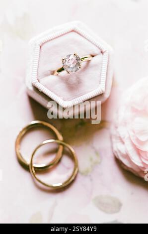 Der zierliche Verlobungsring sitzt auf einem weichen Samtkissen, begleitet von zwei goldenen Eheringen auf einer Blumenoberfläche. Stockfoto