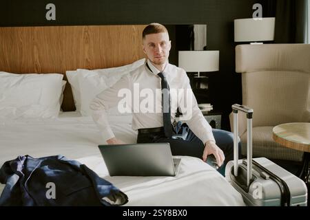 Hochwinkel-Porträt eines erfolgreichen Geschäftsmannes, der auf dem Bett sitzt, mit Laptop und Blick auf die Kamera, seinem Jack auf dem Bett und silbernem Koffer in der Nähe des Doppelbetts Stockfoto