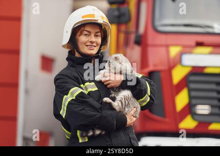Gegen einen großen Truck zu stehen und eine schöne Katze zu halten. Feuerwehrfrau in Uniform arbeitet im Department. Stockfoto