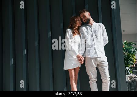 Stehen und halten sich gegenseitig an den Händen. Das schöne junge Paar steht zusammen vor dunkelgrünem Hintergrund. Stockfoto
