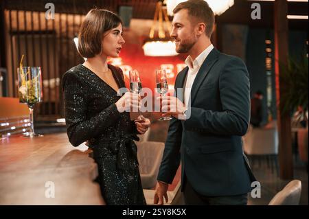 Schönes Paar in schwarzer eleganter Kleidung ist drinnen auf der Party mit Alkohol in den Händen. Stockfoto