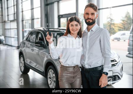 Mit Schlüsseln in der Hand stehen. Glückliche Besitzer. Mann mit Frau in weißer Kleidung ist zusammen im Autohaus. Stockfoto