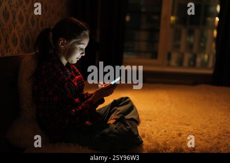 Seitenansicht eines konzentrierten Mädchens, das nachts im dunklen Schlafzimmer auf dem Bett sitzt, von der Bildschirmbeleuchtung beleuchtet, mobile Apps, Spiele und soziale Netzwerke genießt Stockfoto