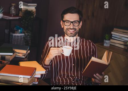 Junger Mann, der einen entspannenden Moment im Haus genießt, ein Buch und eine Tasse Kaffee in der Hand hält, umgeben von Büchern, in einem gemütlichen und stilvollen Wohnraum. Stockfoto