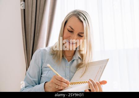 Lächelndes Mädchen, das mit einem Stift in einem Notizbuch schreibt, 35-37 Jahre alt, europäisches Aussehen, Nahaufnahme, mit blauem Hemd. Hochwertige Fotos Stockfoto