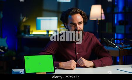Technikingenieur präsentiert ein Tablet mit grünem Bildschirm in einer preisgünstigen Preisklasse und filmt aus einem schwach beleuchteten Heimstudio. Content Creator überprüft das digitale Modell vor Online-Publikum, Kamera A Stockfoto