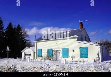 Ein traditionelles Tafel-Haus mit hellen türkisfarbenen Rollläden und einem weißen Pfahlzaun ist umgeben von tiefen Schneebänken unter einem klaren blauen Winterhimmel. Stockfoto