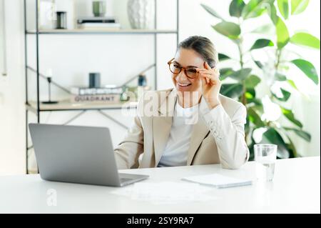 Fröhliche erfolgreiche hübsche kaukasische Geschäftsfrau in einem stilvollen eleganten Anzug und einer Brille, sitzt an ihrem Arbeitsplatz mit Laptop in einem modernen Büro, schaut auf den Bildschirm und lächelt glücklich Stockfoto