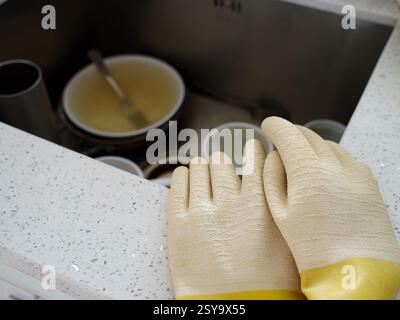 Gelbe und beige Gummihandschuhe am Küchentisch über schmutzigem Geschirr im Waschbecken Stockfoto