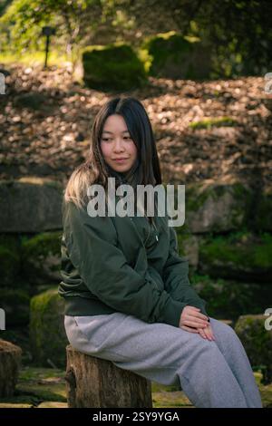 Die junge Frau in lässiger Kleidung sitzt friedlich auf einem Holzhocker in einer natürlichen Gartenumgebung, umgeben von moosigen Steinen und sanftem Licht. Golden Gate Stockfoto