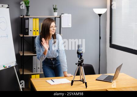 Positive Bloggerin, die auf einem Stativ an der Smartphone-Webcam spricht, einen Beitrag für den Vlog aufnimmt. Online-Lehrer, Coach, Webinar zu Hause. Pressen Stockfoto
