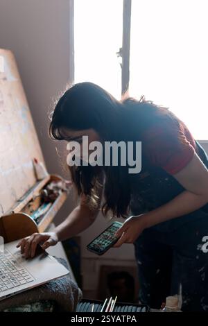 Eine junge Lateinfrau konzentriert sich auf das Bemalen einer Leinwand in ihrem gemütlichen Atelier. Ihr Arbeitsbereich ist voller Pinsel, Farben und ihrer Kreativität. Stockfoto