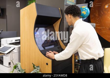 Ein Mitarbeiter spielt während einer Pause in einem modernen Büro auf einem Spielautomaten im Retro-Stil. Zu den umliegenden Gegenständen gehören ein Globus, ein Drucker und Wandplakate, Stockfoto