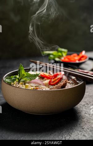 Frisch zubereitetes vietnamesisches Rindfleisch Pho Nudelsuppe (Pho Bo) in einer Keramikschale, mit Dampf, der über Garnituren von Basilikum, Zwiebeln, Chili und Limette steigt, wi Stockfoto